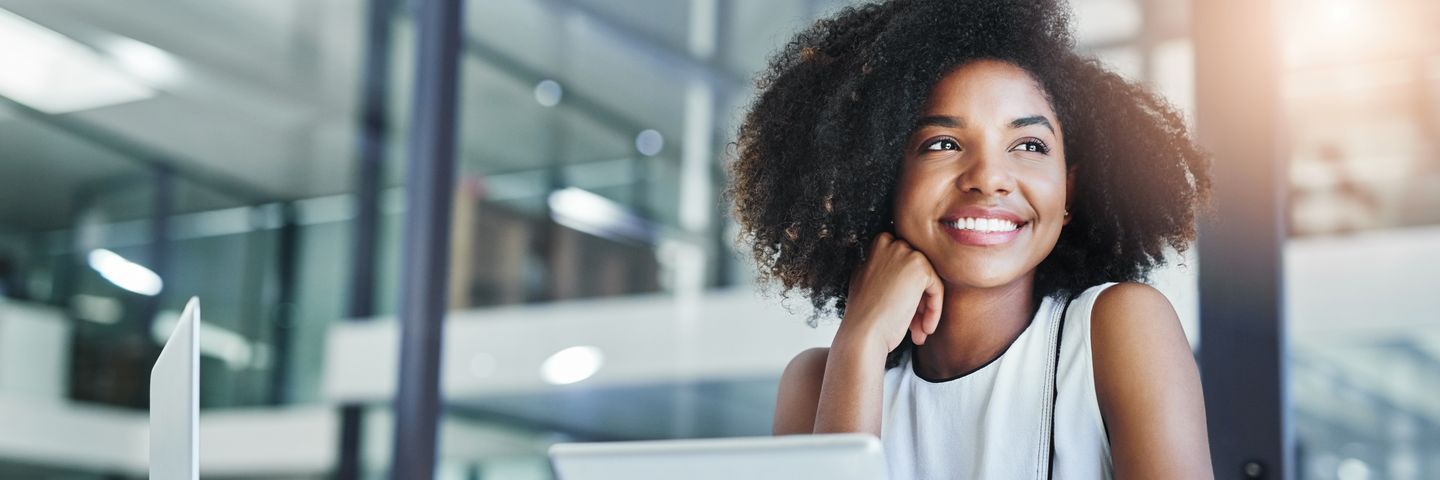 A joyful african american woman with curly hair sits at her desk in a modern office, smiling dreamily while leaning on her hand, with a laptop in front of her.