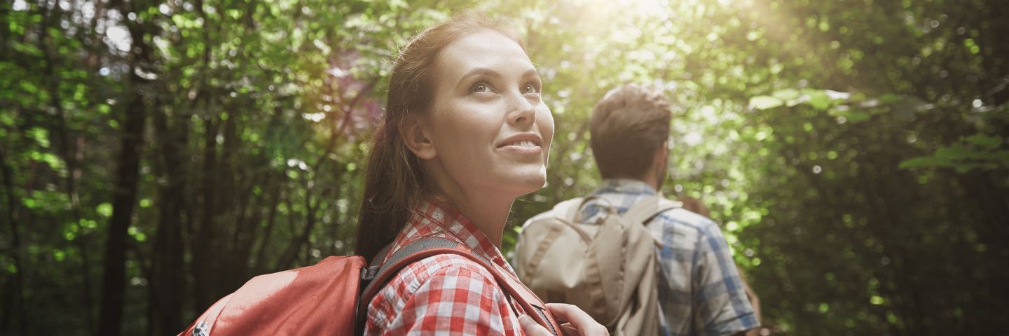 A woman with a backpack looks up happily in a sunlit forest with a male companion walking ahead.