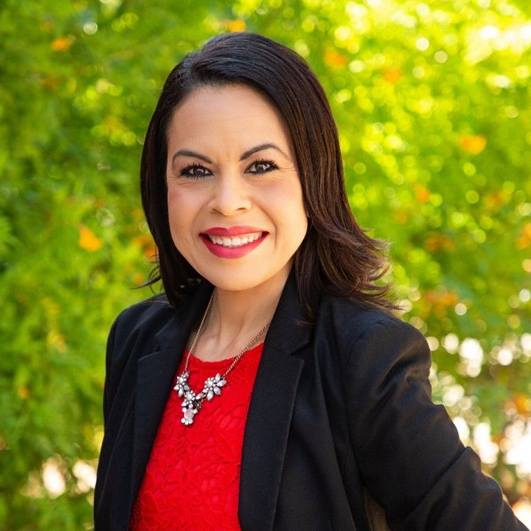 A professional portrait of a smiling woman with medium-length dark hair, wearing a red blouse, black blazer, and a statement necklace, with lush green trees blurred in the background.