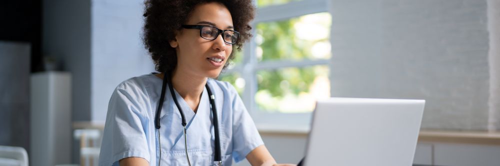 A female healthcare professional wearing glasses and a stethoscope is working on a laptop in a well-lit office.