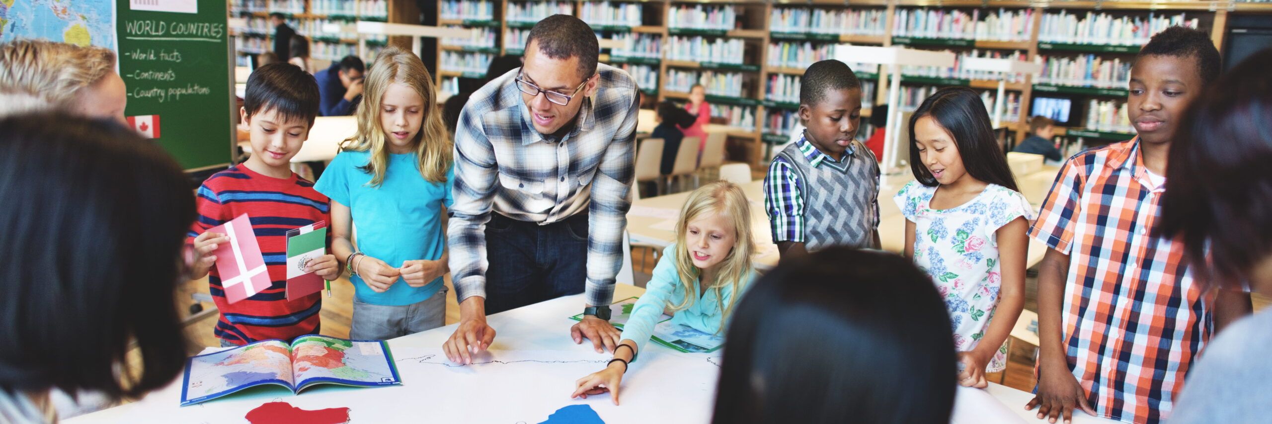 A diverse group of elementary students with a teacher in a school library, engaged in a classroom activity with books and flags on the table, similar to practices taught in the best online master's programs.