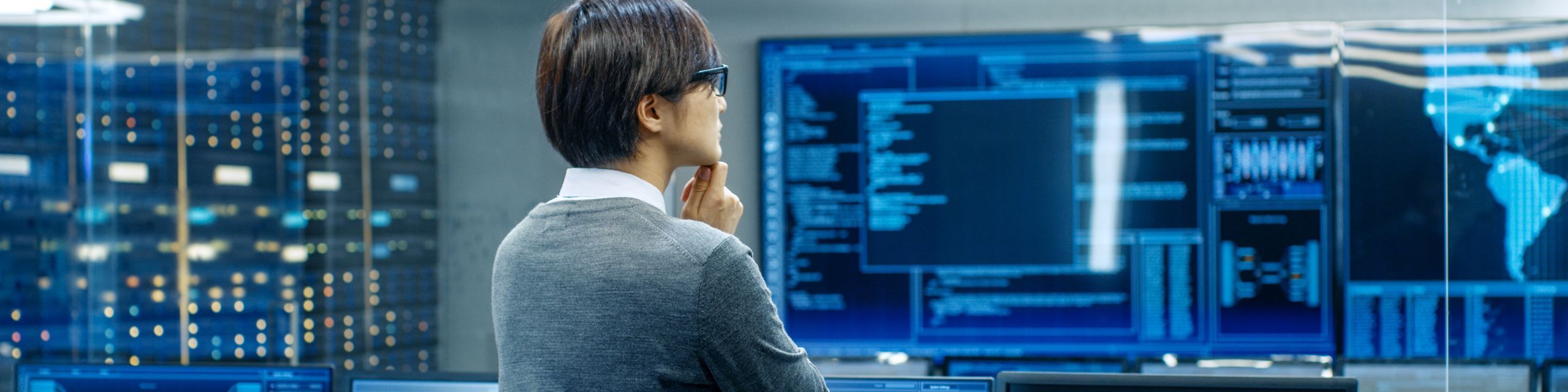 A person wearing glasses stands in a server room, looking at a large screen displaying various code and data visualizations. The room is filled with computer equipment, indicating a high-tech and secure environment.
