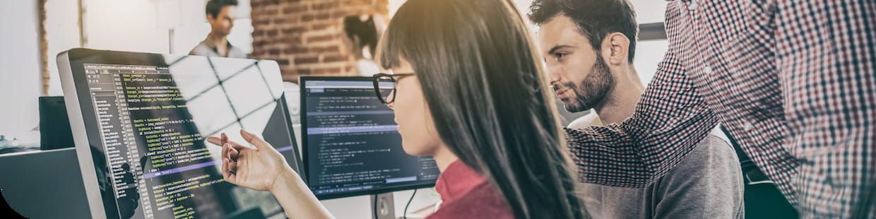 A young women with dark hair and glasses points to code on a computer monitor while a man looks over her shoulder.