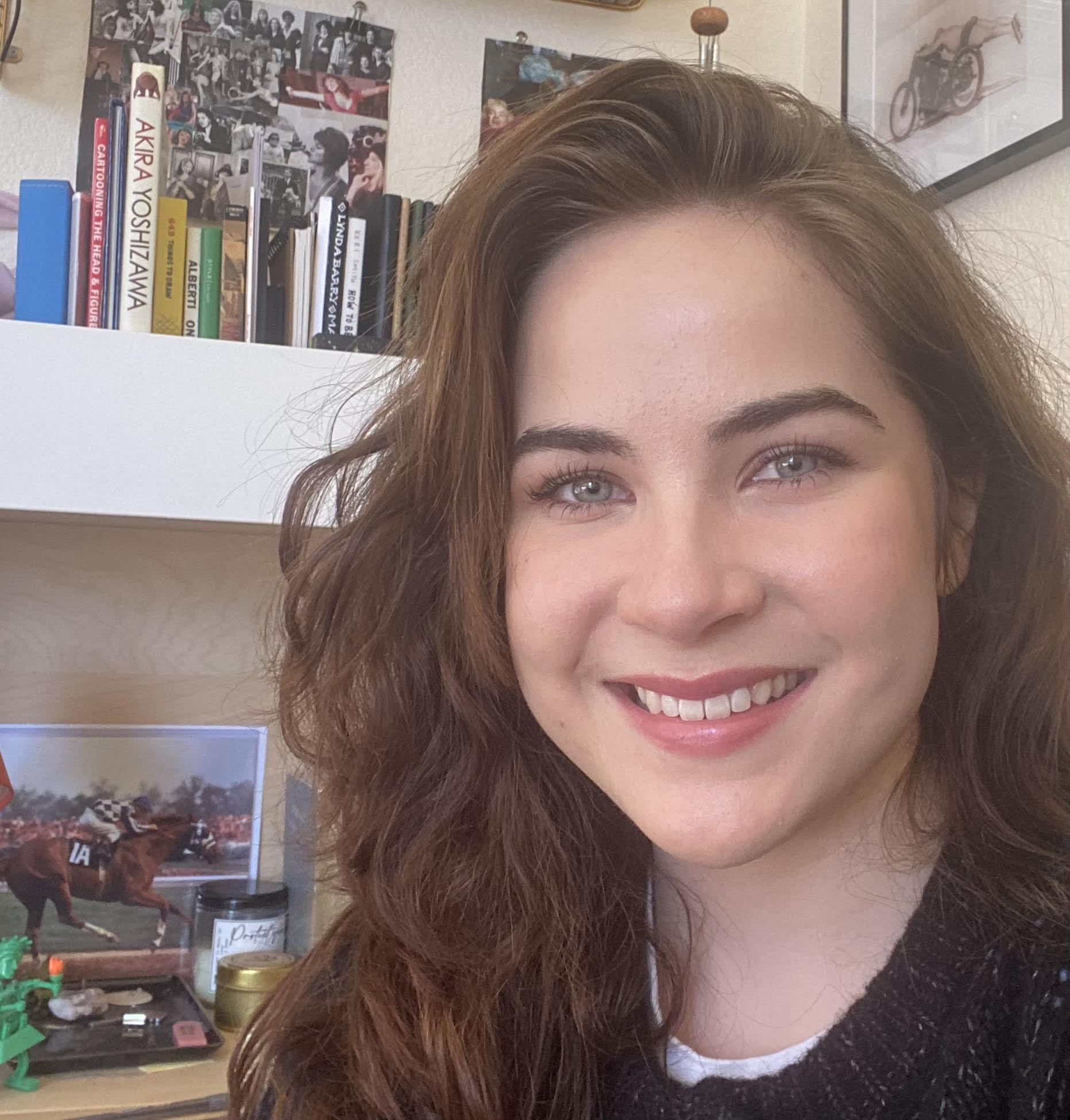 A young woman with long brown hair smiles at the camera, sitting indoors with books, photos, and a framed picture of a horse behind her. The setting appears to be a cozy, personal workspace or study.