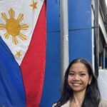 A woman with long dark hair smiles in front of a large Philippine flag displayed vertically against a blue building.