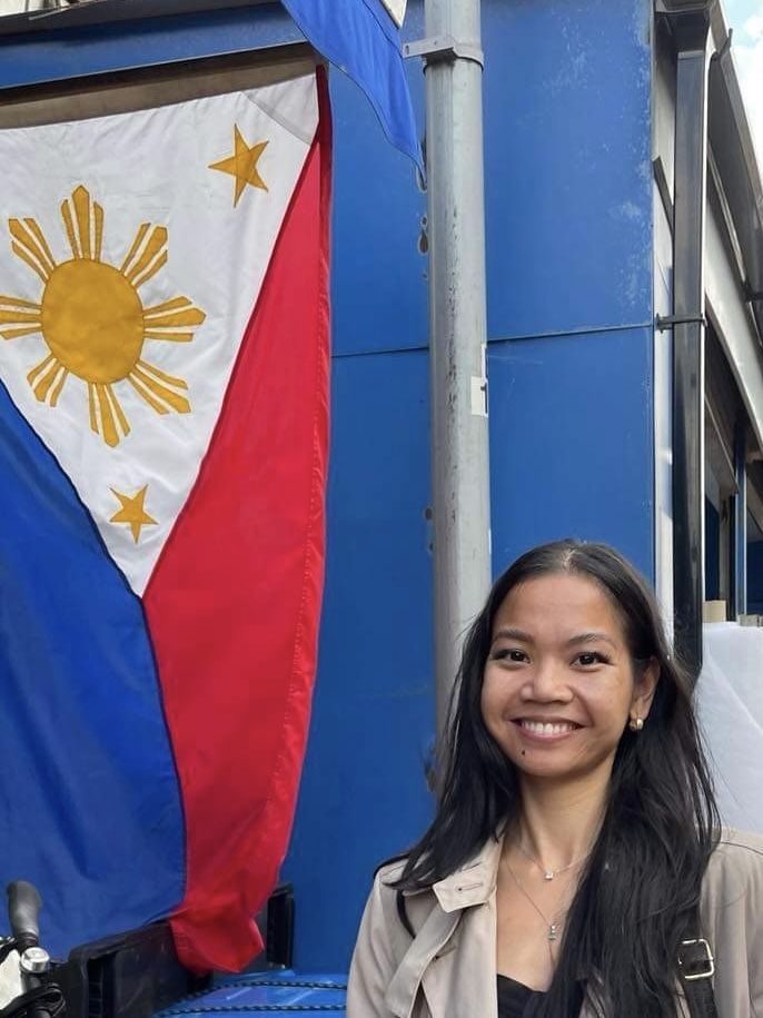A woman with long dark hair smiles in front of a large Philippine flag displayed vertically against a blue building.