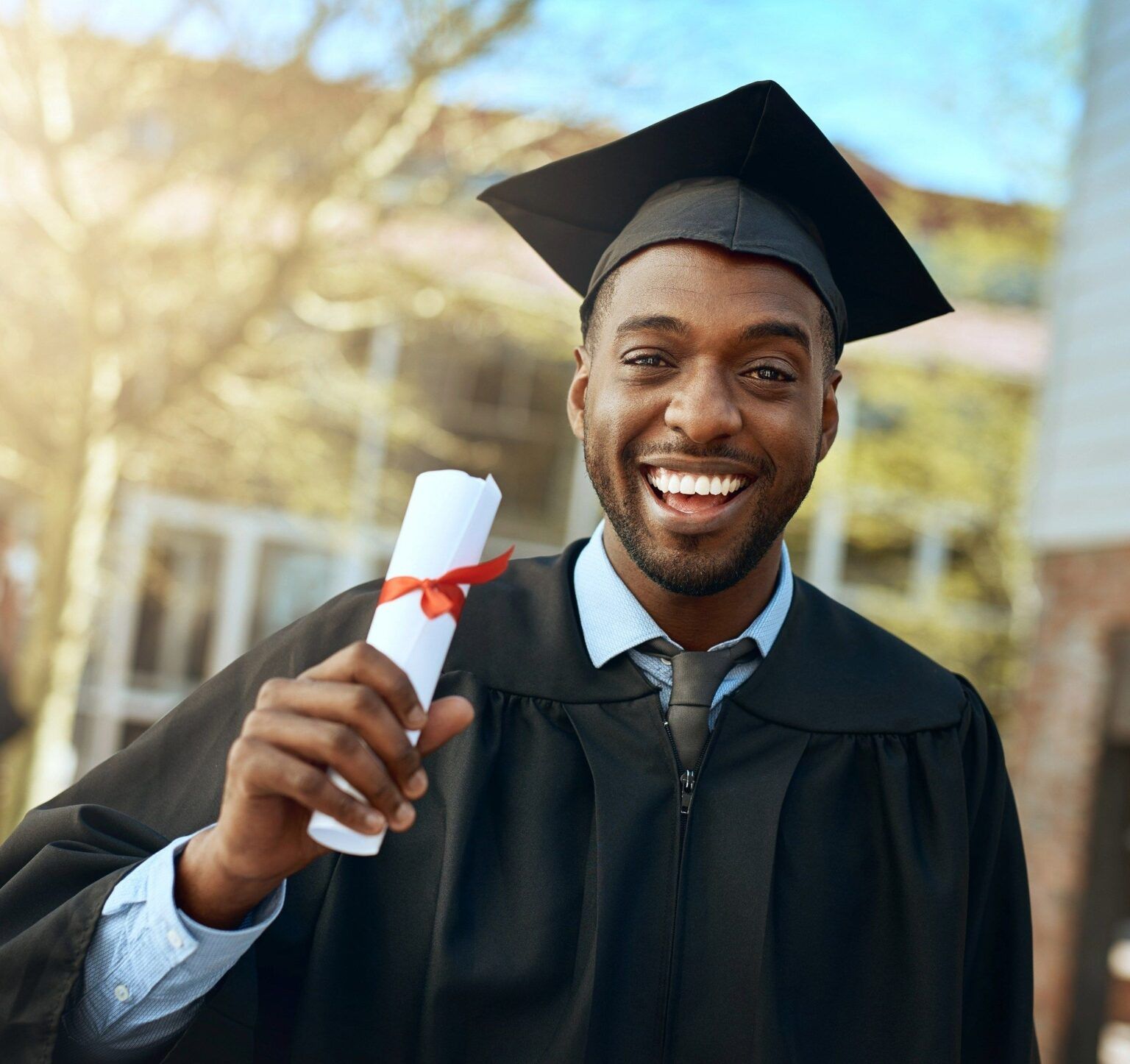A young man wearing a graduation cap and gown smiles while holding a diploma tied with a red ribbon, standing outdoors with blurred buildings and trees in the background.