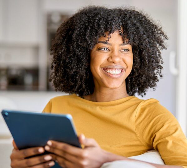 A smiling woman with curly hair in a yellow top sits indoors, holding a tablet. She looks to the side, appearing happy and relaxed in a bright, modern living space.