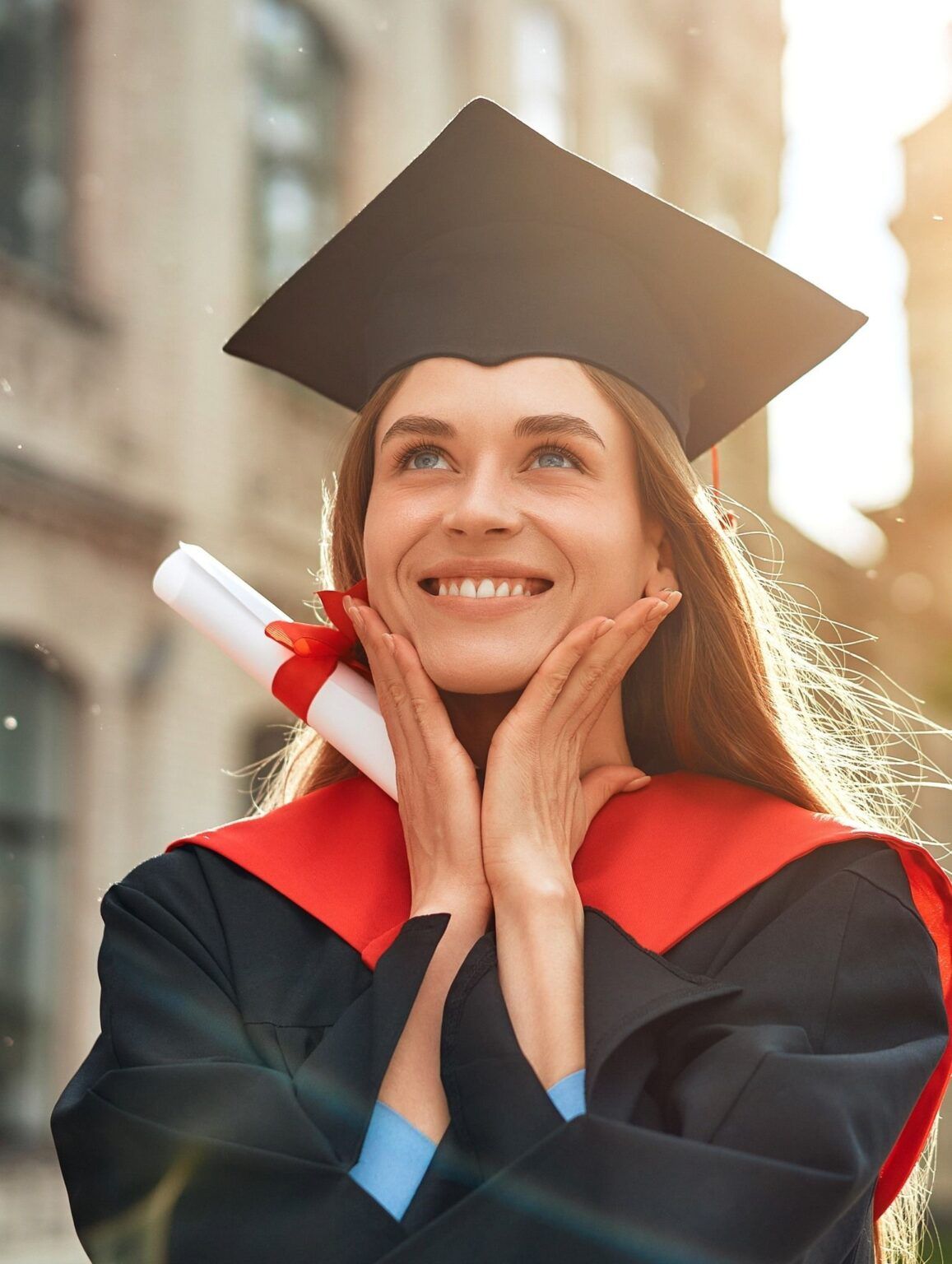 A young woman in a graduation cap and gown smiles joyfully, holding her diploma and touching her face with both hands. Sunlight shines behind her, creating a warm and celebratory atmosphere.