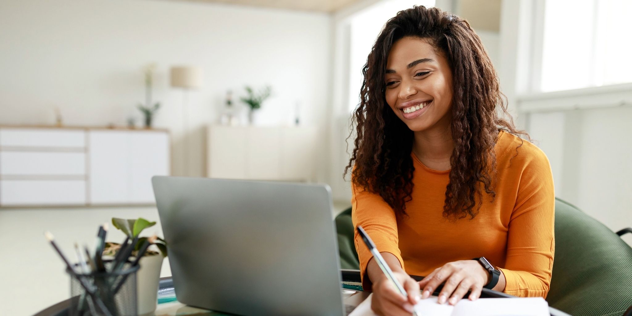 A woman with long curly hair wearing an orange shirt sits at a table, smiling while writing in a notebook and looking at an open laptop in a bright, modern room.
