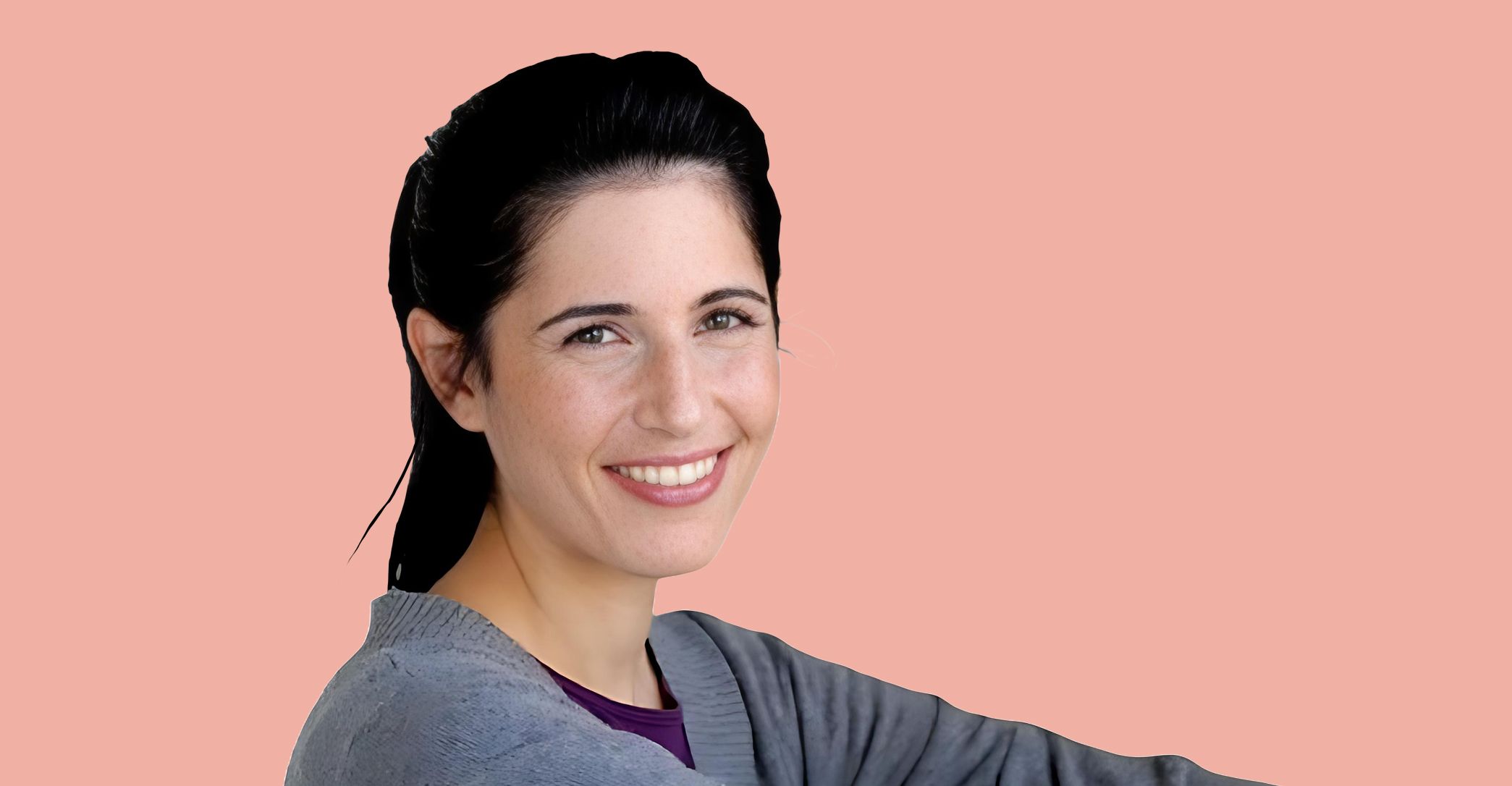 A woman with dark hair and a gray sweater smiles while looking at the camera, set against a plain light pink background.