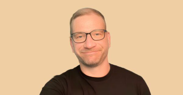 A smiling man with short light hair, glasses, and a beard wearing a black t-shirt, standing in front of a solid beige background.