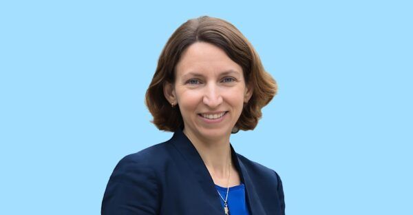 A woman with short brown hair, wearing a navy blazer over a blue top, smiles at the camera against a light blue background.