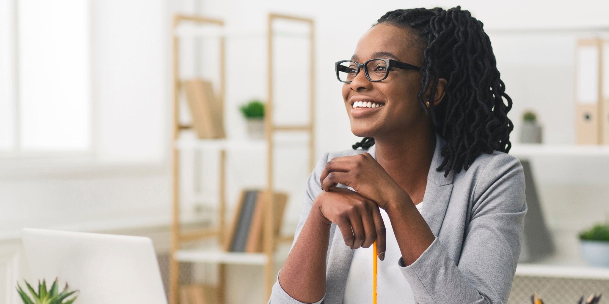 A woman wearing glasses and a gray blazer smiles while sitting at a desk in a bright office, holding a pencil. Shelves with books and plants are visible in the background.
