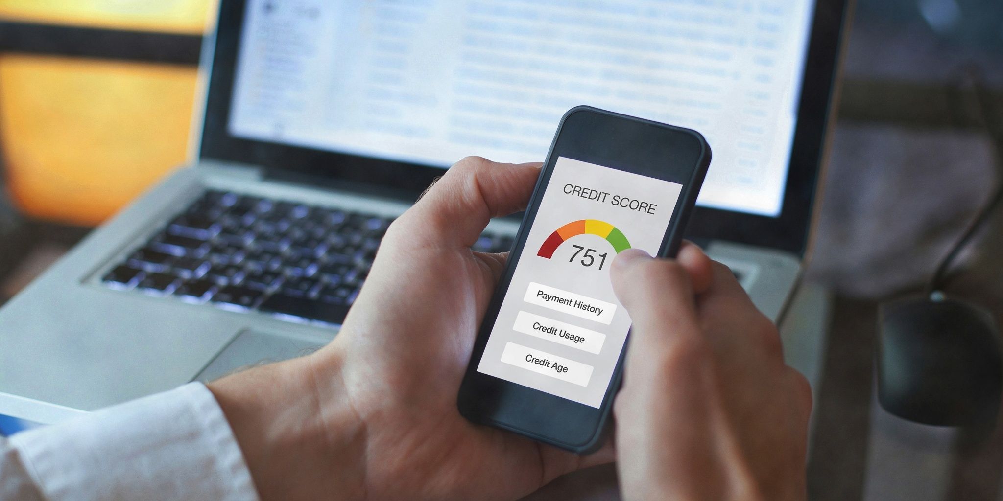 A person holds a smartphone displaying a credit score of 751, with colored gauge and categories like payment history, credit usage, and credit age. A laptop and a computer mouse are visible in the background.