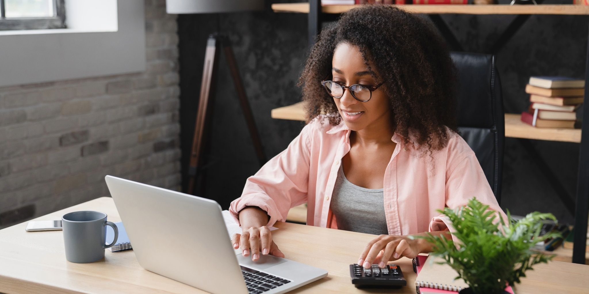A woman with curly hair and glasses sits at a desk using a laptop and a calculator. There is a blue mug, a notebook, a plant, and shelves with books in the background. She appears to be working or studying.