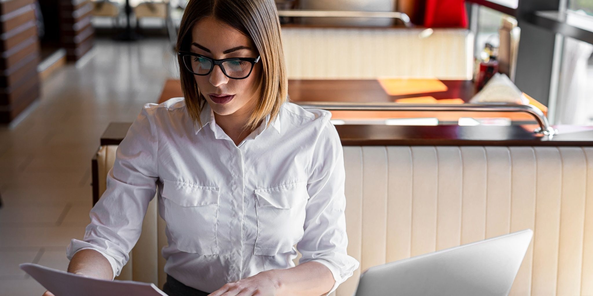 A woman with straight, shoulder-length hair and glasses sits in a booth at a modern restaurant, focused on reading a document while using a laptop. She wears a white button-up shirt and appears to be working.