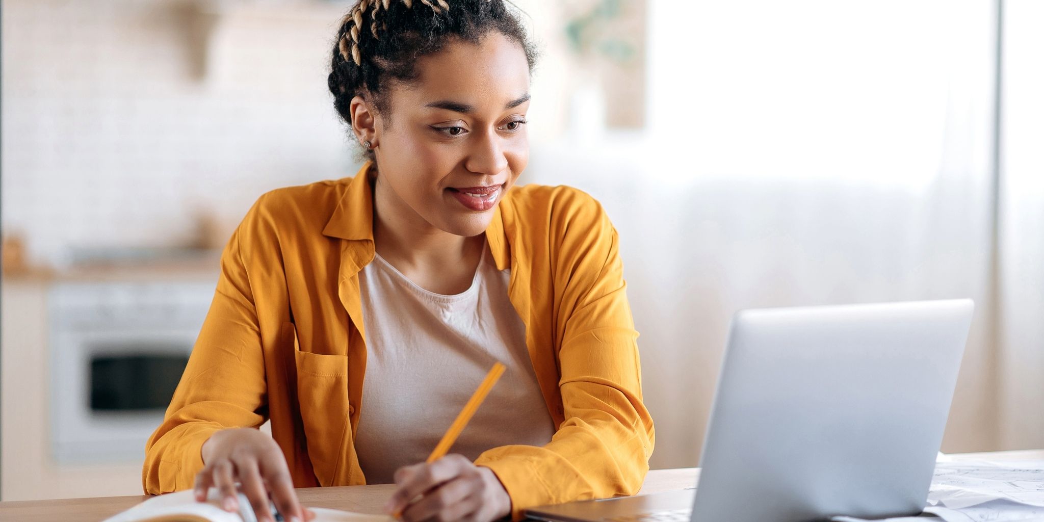 A woman wearing a yellow shirt sits at a table, smiling while looking at a laptop screen. She holds a pencil and writes in a notebook, appearing focused and engaged in her work.