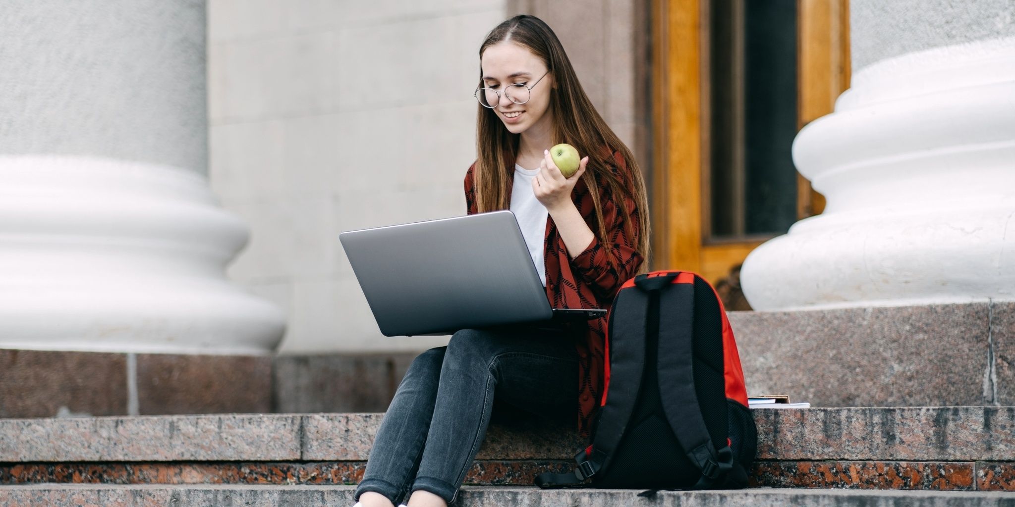 A young woman with glasses sits on stone steps outside, holding an apple and using a laptop. A red and black backpack is next to her. She looks focused and relaxed.