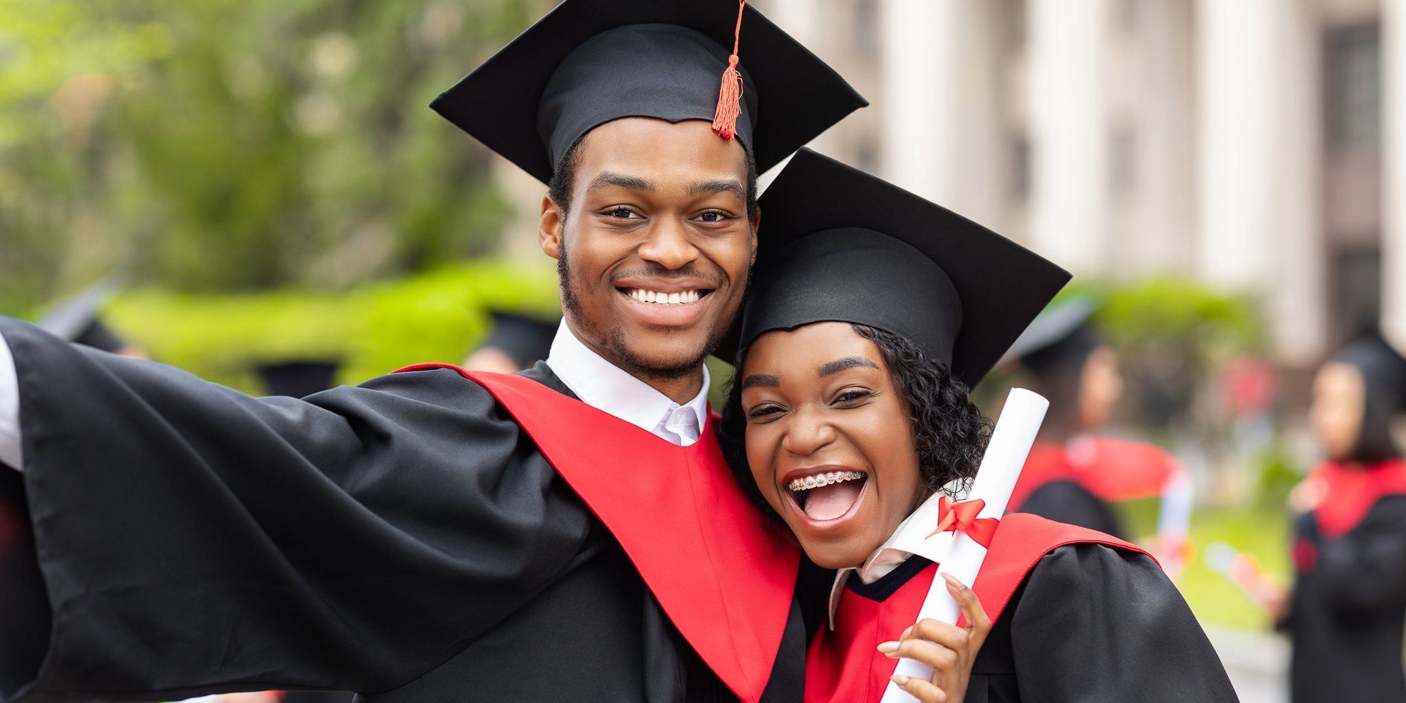 Two graduates in caps and gowns smile and pose for a selfie outdoors. One holds a diploma, and both wear black gowns with red stoles. There are other graduates and greenery in the background.