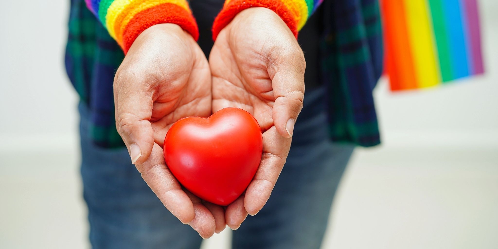 A person wearing a rainbow-striped sleeve holds a red heart-shaped object in their cupped hands. A rainbow flag is visible in the background, symbolizing LGBTQ+ pride and love.