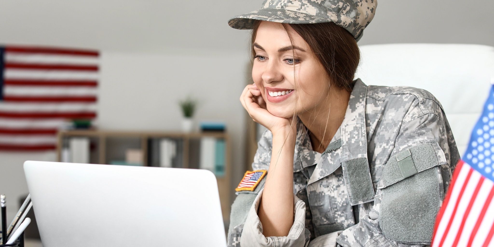 A smiling woman in military uniform sits at a desk, looking at a laptop. An American flag patch is on her sleeve, and a small U.S. flag is on the desk. A blurred flag hangs in the background.