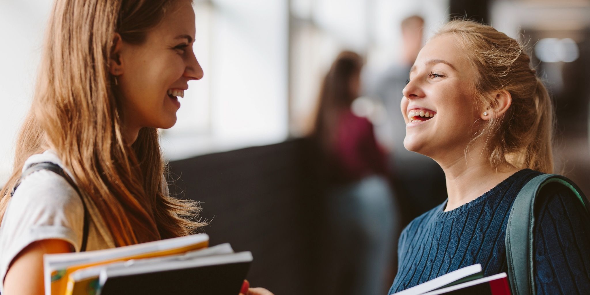 Two smiling young women with long hair stand in a hallway, holding books and backpacks, engaged in a cheerful conversation. The background is softly blurred, showing other people and windows.