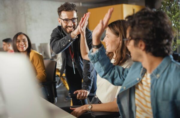 Four coworkers are smiling in a modern office. Two people in the foreground are giving a high-five, while two others look on, also smiling, suggesting a moment of workplace celebration or teamwork.