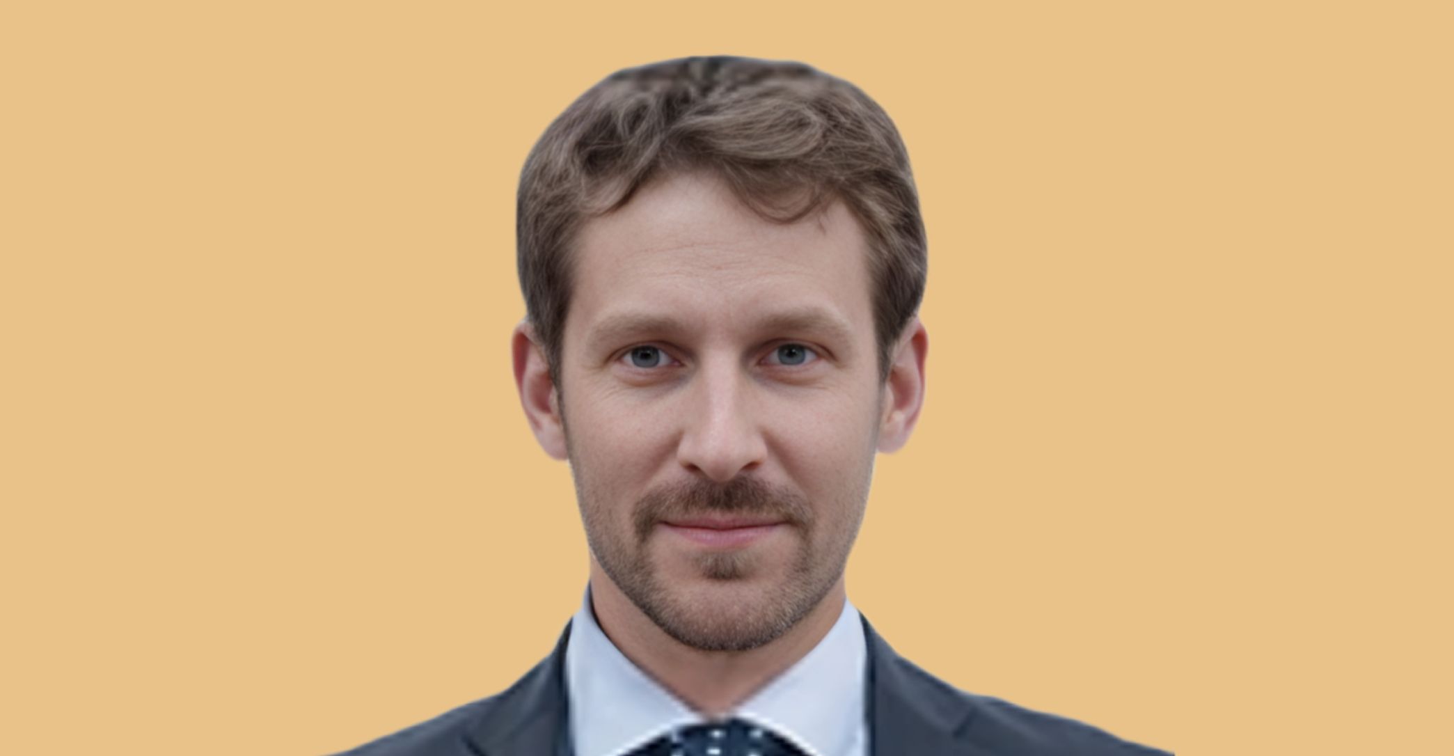 A man with light brown hair, trimmed beard, and blue eyes, wearing a dark suit, white shirt, and polka-dot tie, standing against a plain beige background.
