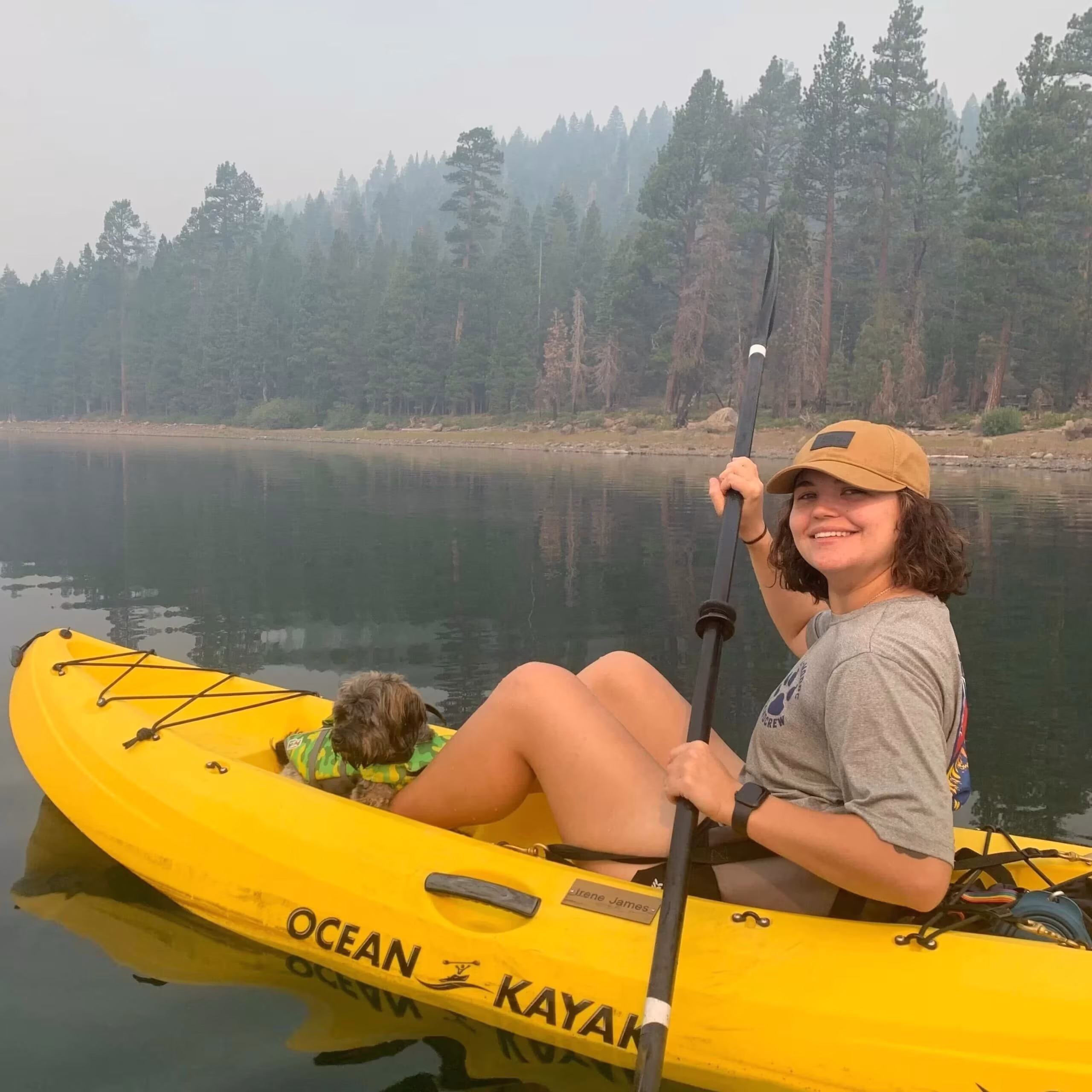 A woman smiles while sitting in a yellow kayak on a calm lake, holding a paddle. A small dog sits in the kayak with her. Tall pine trees and misty mountains are visible in the background.