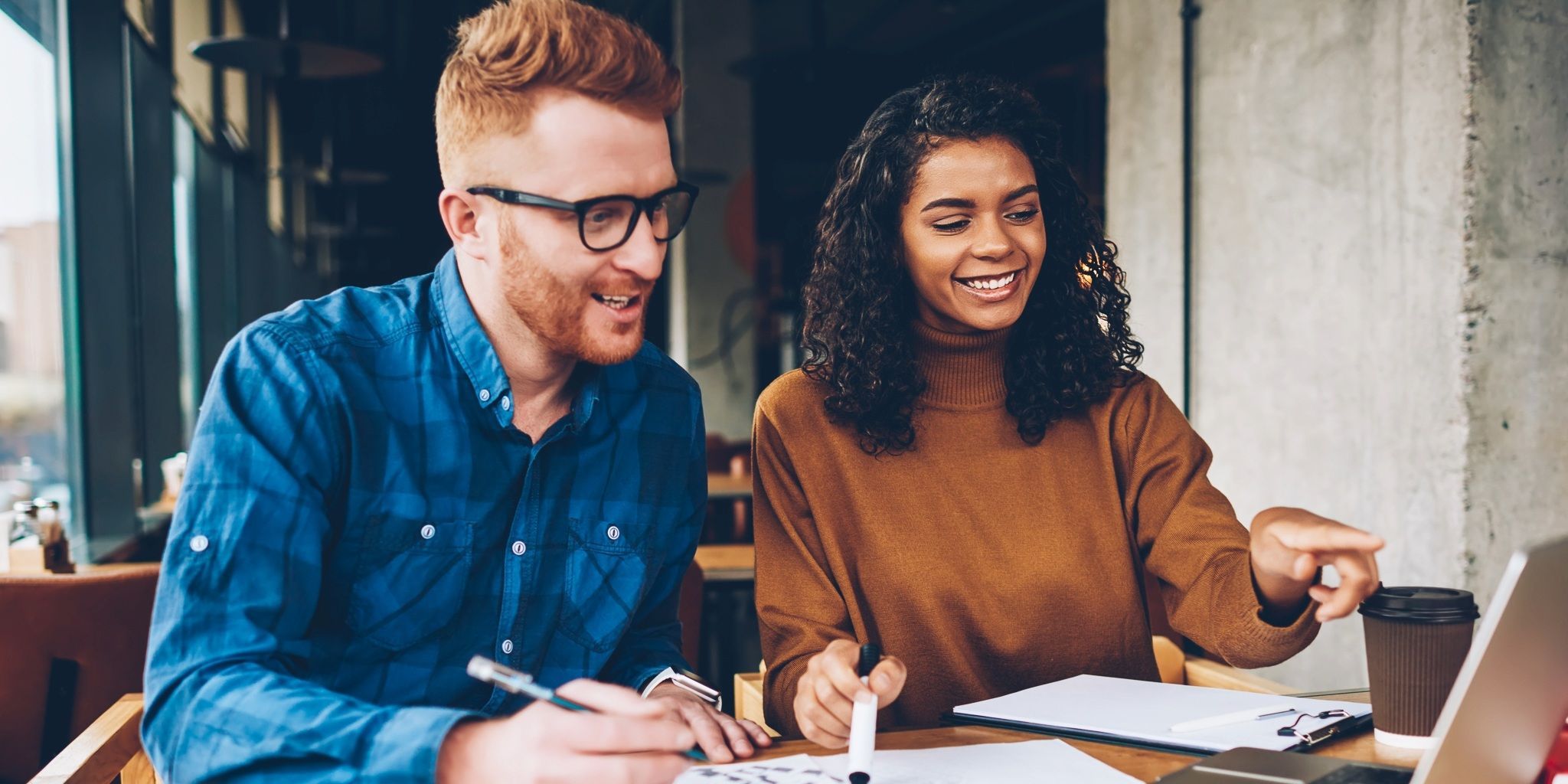 Two people sit at a table with papers and a laptop, smiling and looking at the laptop screen. One person points at the screen while the other takes notes. They appear to be working together in a modern café.
