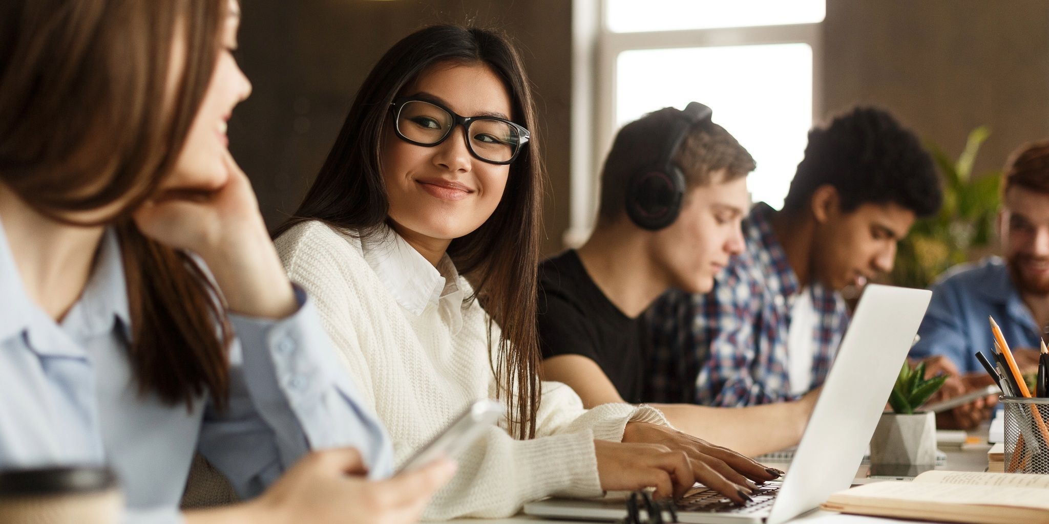 A group of young adults sit at a table working on laptops and writing. One woman in glasses smiles at another, while others focus on their devices and notebooks. The atmosphere appears casual and collaborative.