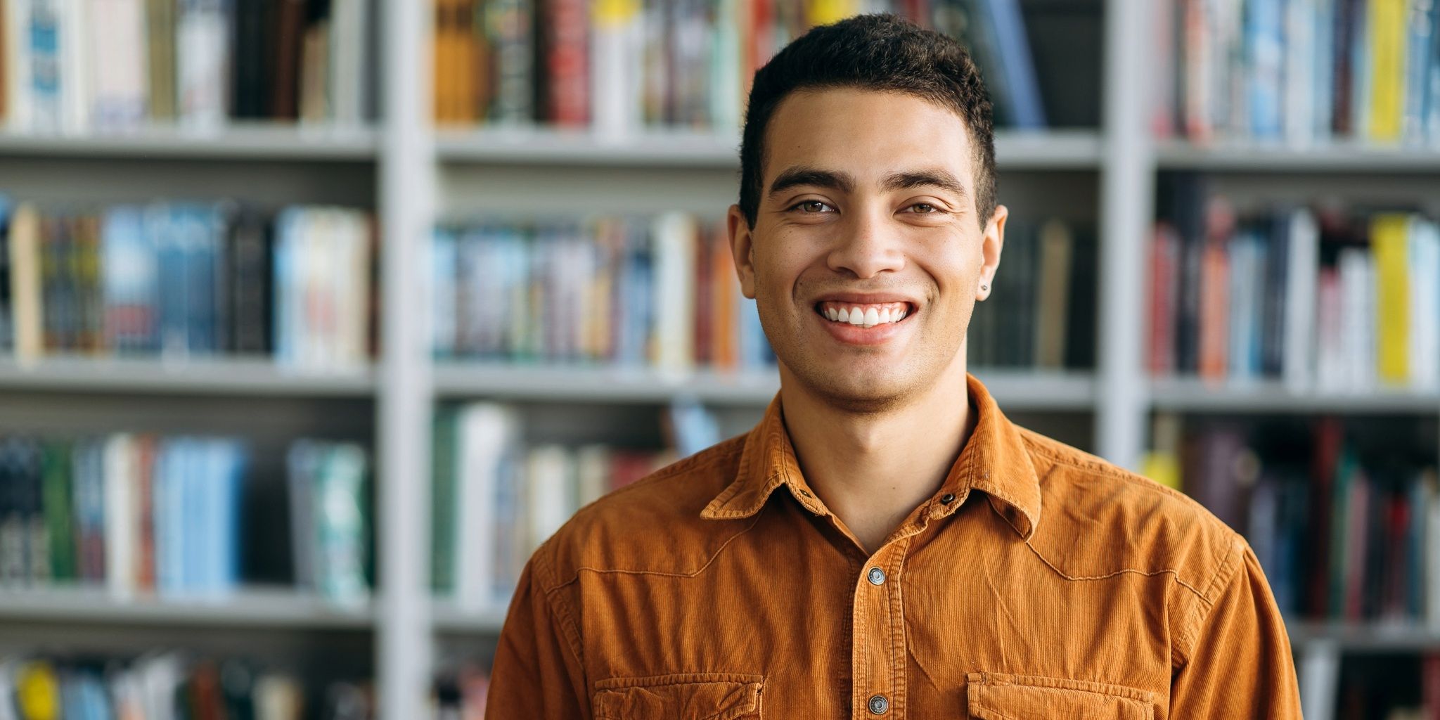 A young man wearing a brown button-up shirt smiles at the camera while standing in front of a bookshelf filled with books.