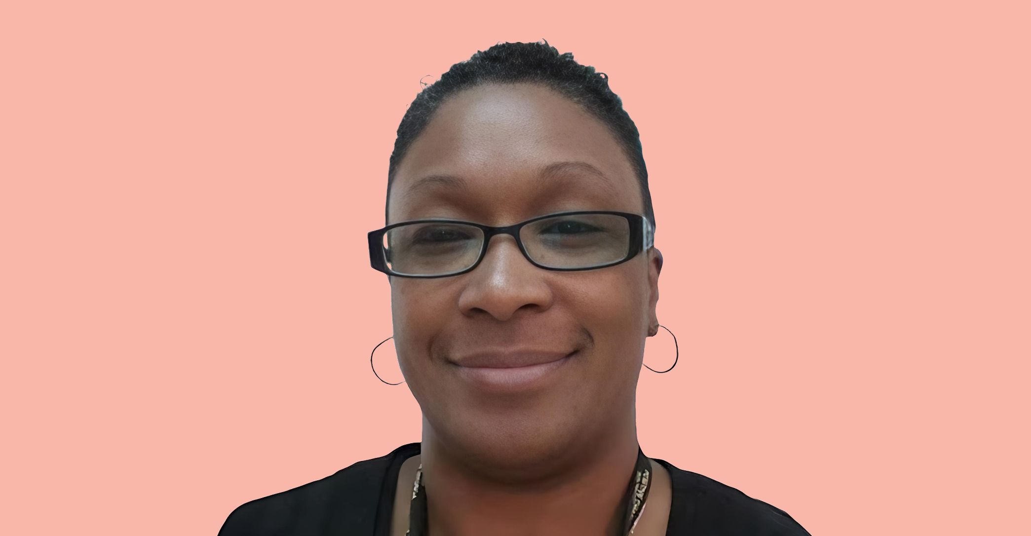 A person with short hair, wearing glasses and hoop earrings, smiles at the camera against a solid light pink background.