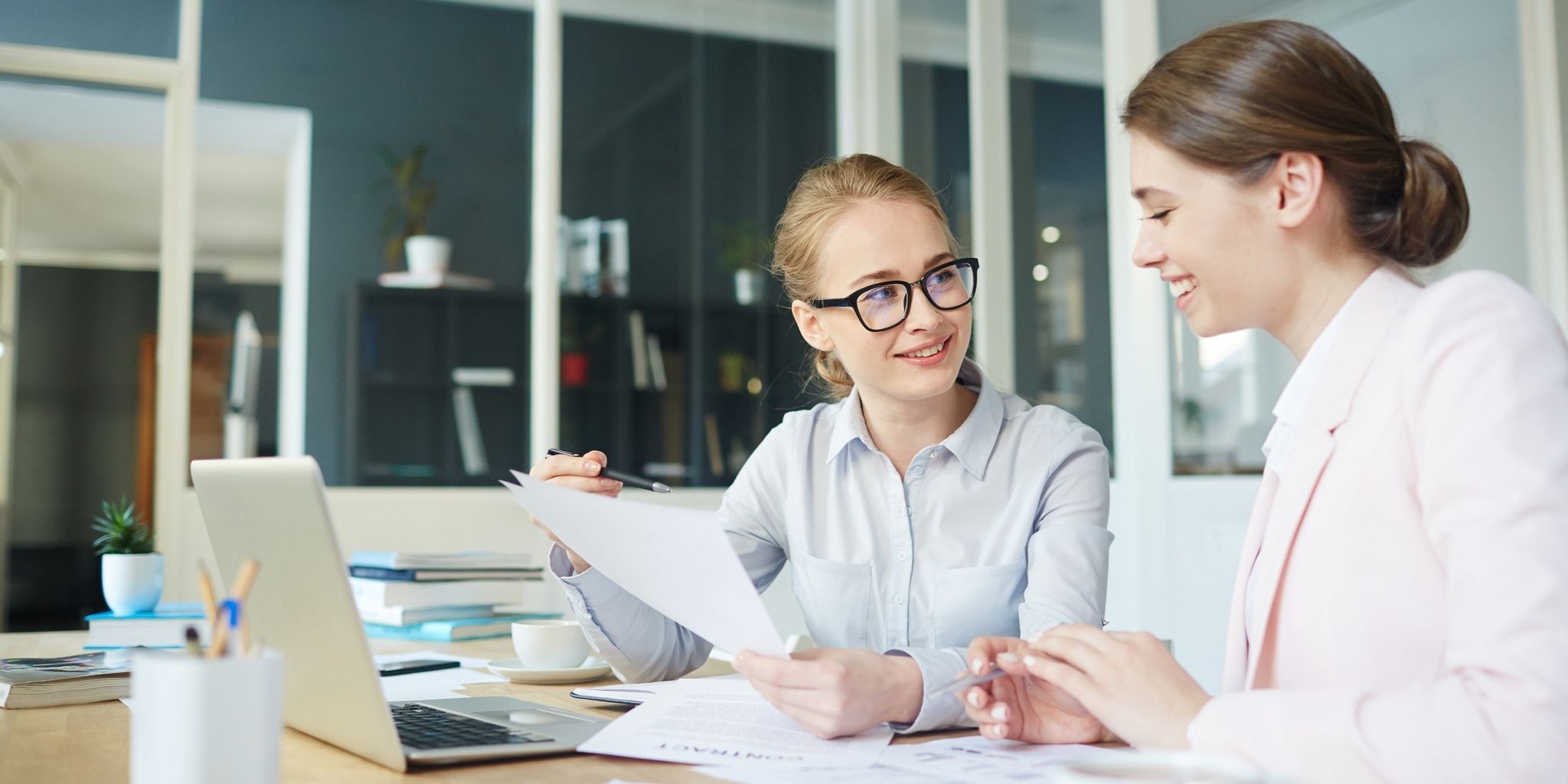 Two women sit at a desk in an office, smiling and discussing documents. One woman holds a sheet of paper and a pen, while the other listens. A laptop and paperwork are on the desk in front of them.