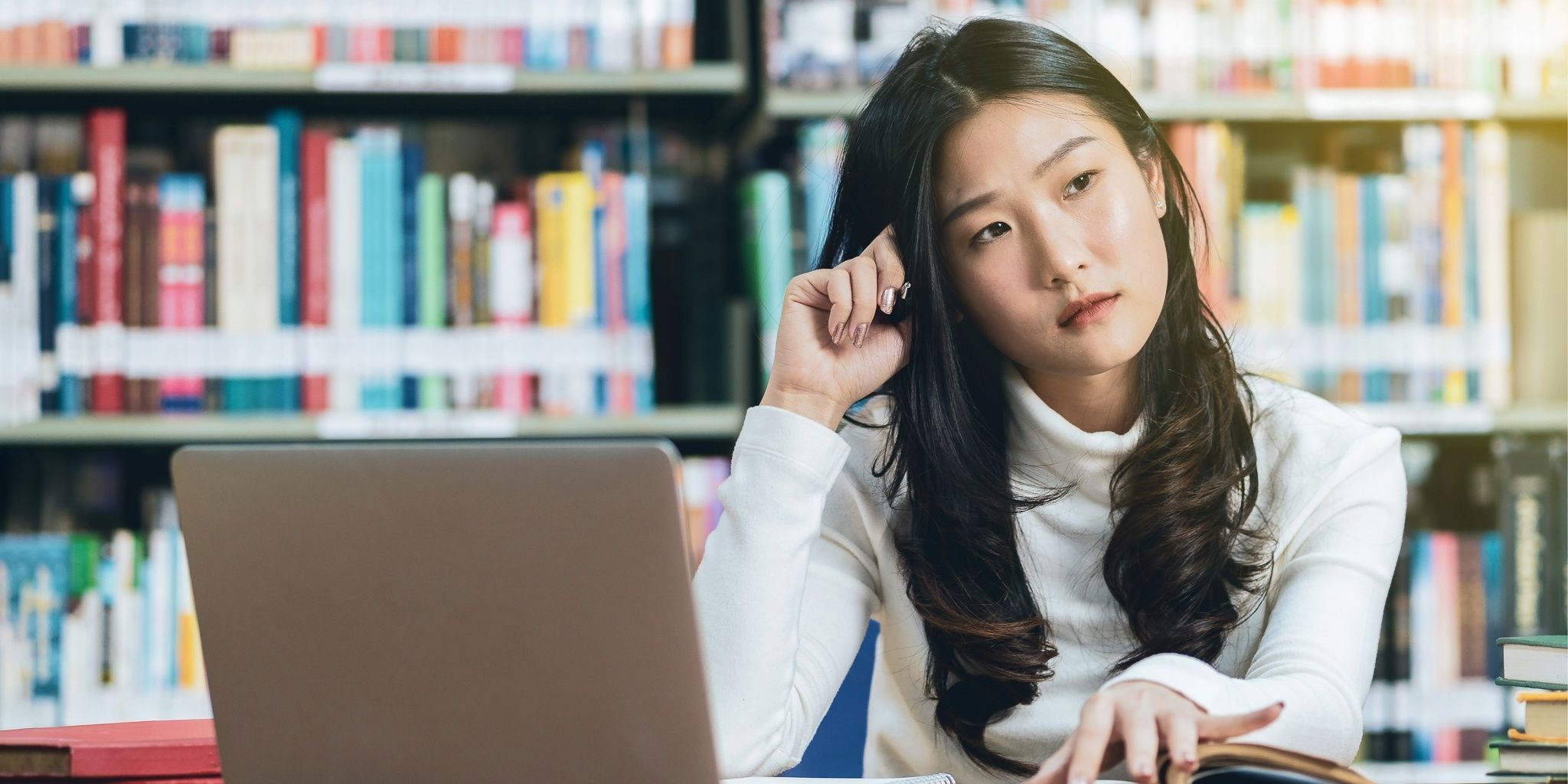 A young woman sits at a desk in a library with an open laptop and book, resting her head on her hand and looking thoughtful or distracted. Bookshelves are visible in the background.