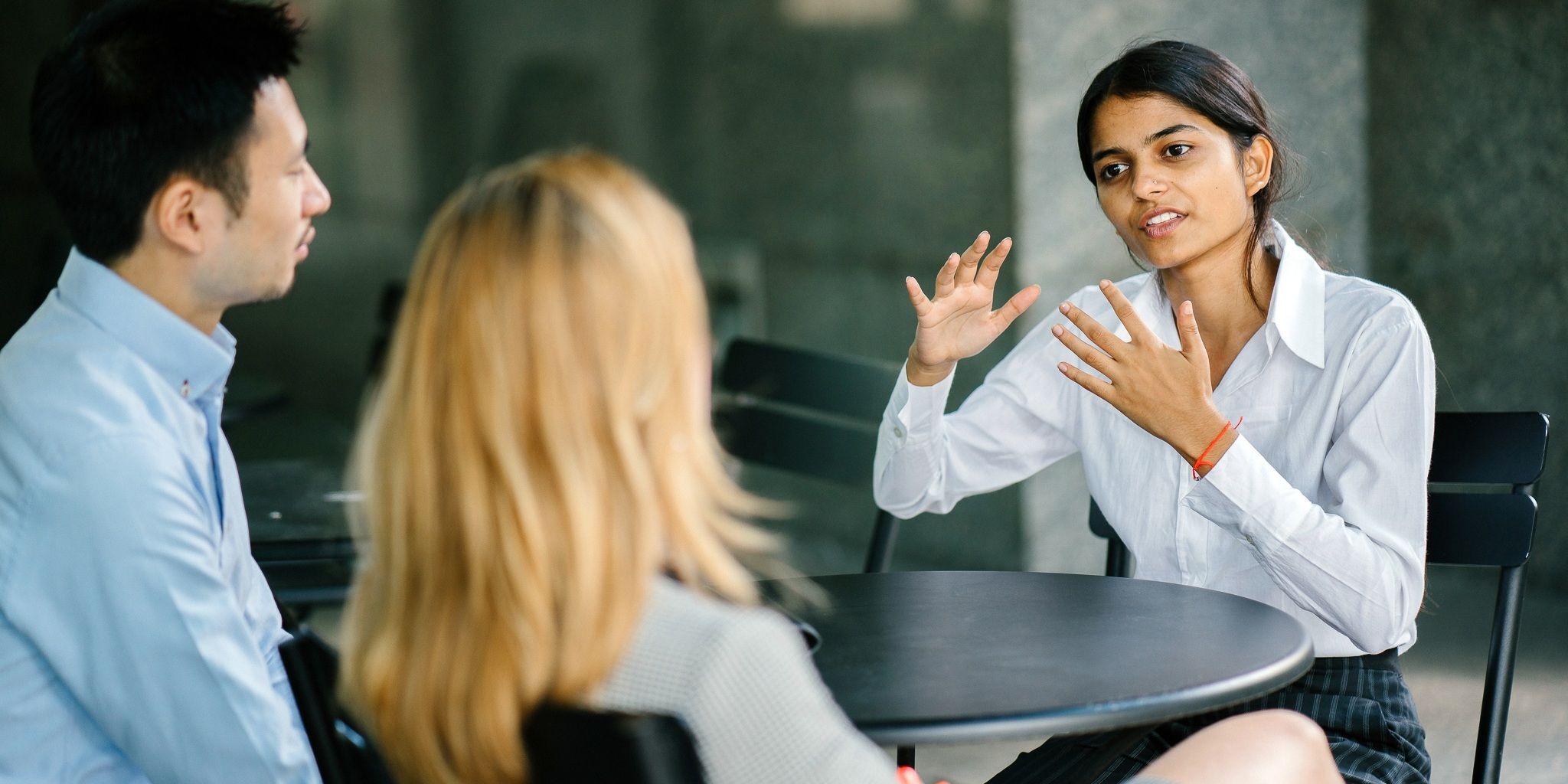 A woman gestures while speaking to two colleagues at a round table during a discussion in a modern indoor setting.