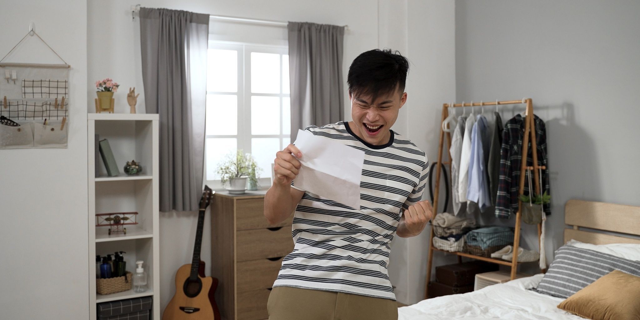 A young man stands in a bedroom, happily reading a piece of paper and clenching his fist in excitement. The room has a guitar, clothes rack, and sunlight coming through the window.