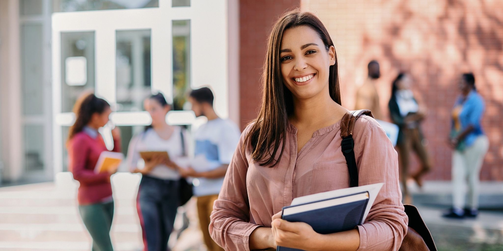 A young woman smiling and holding books stands in the foreground, while a group of students converse and walk in the background outside a school or college building.