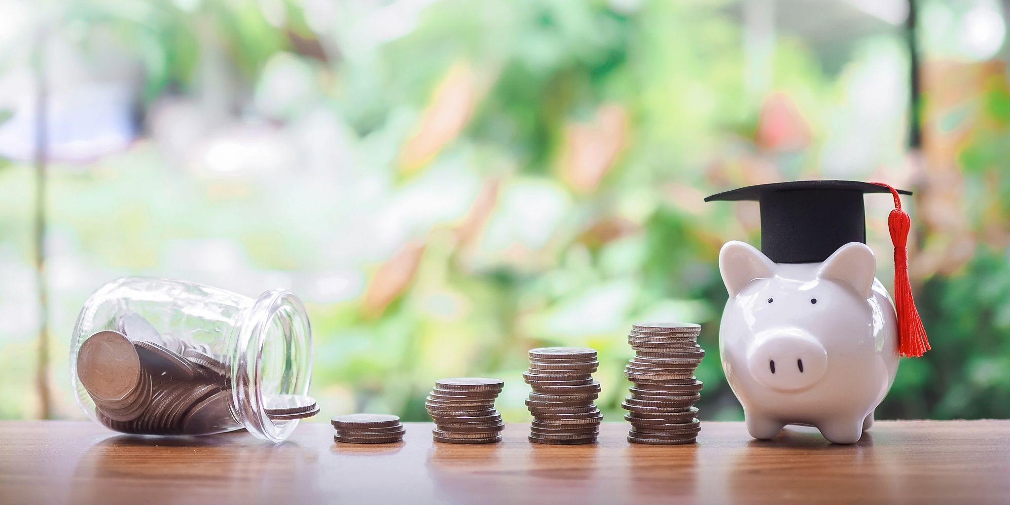 A piggy bank wearing a graduation cap sits beside stacks of coins and a tipped-over jar of coins on a wooden table with a blurred green background.