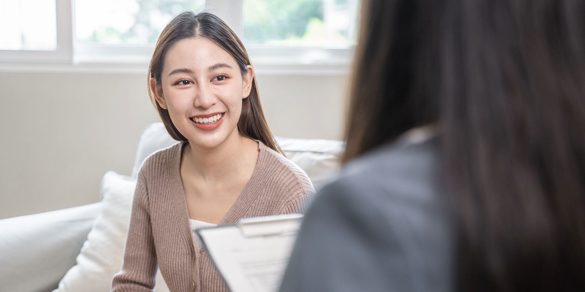 A young woman sits on a couch smiling and talking to another person who is holding a clipboard, suggesting a friendly conversation or counseling session in a bright room.