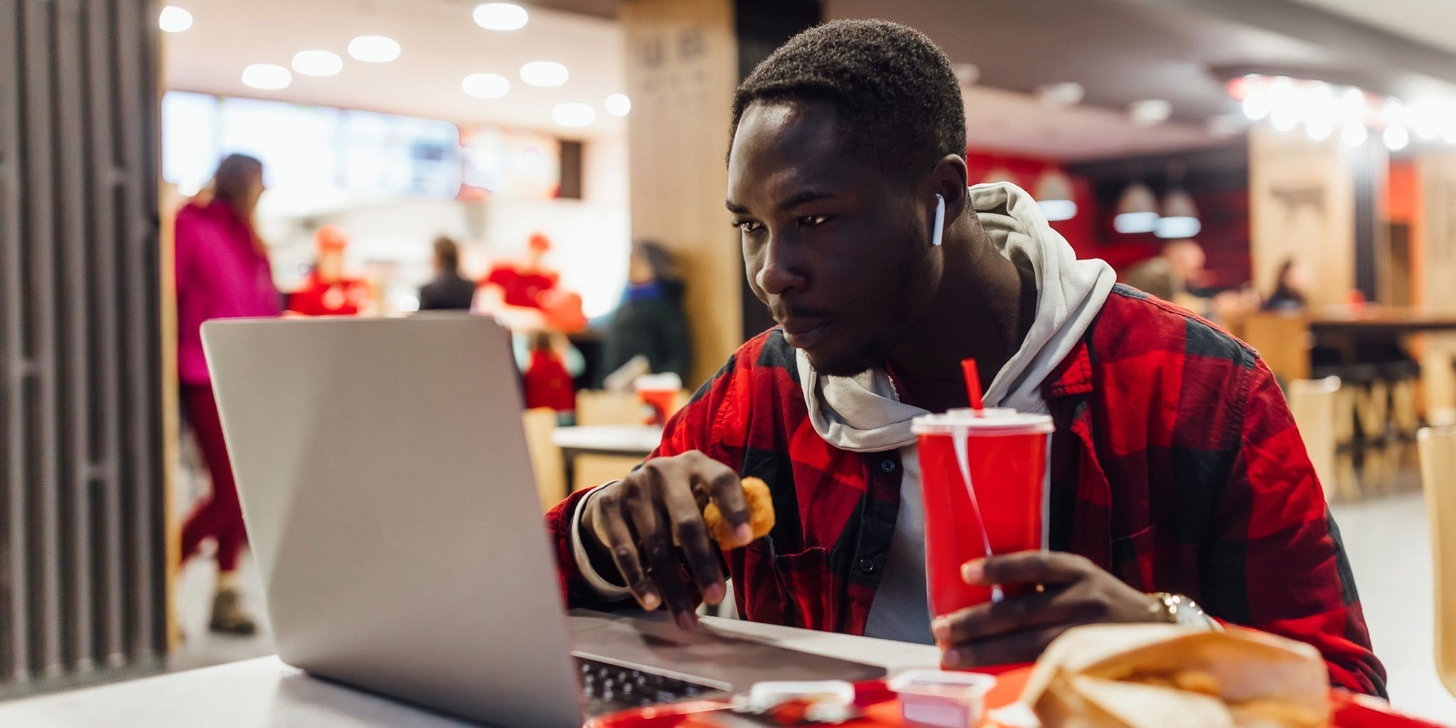 A man wearing a red plaid shirt and wireless earbuds works on a laptop at a fast food restaurant, holding a soft drink and some food, with trays and wrappers on the table.