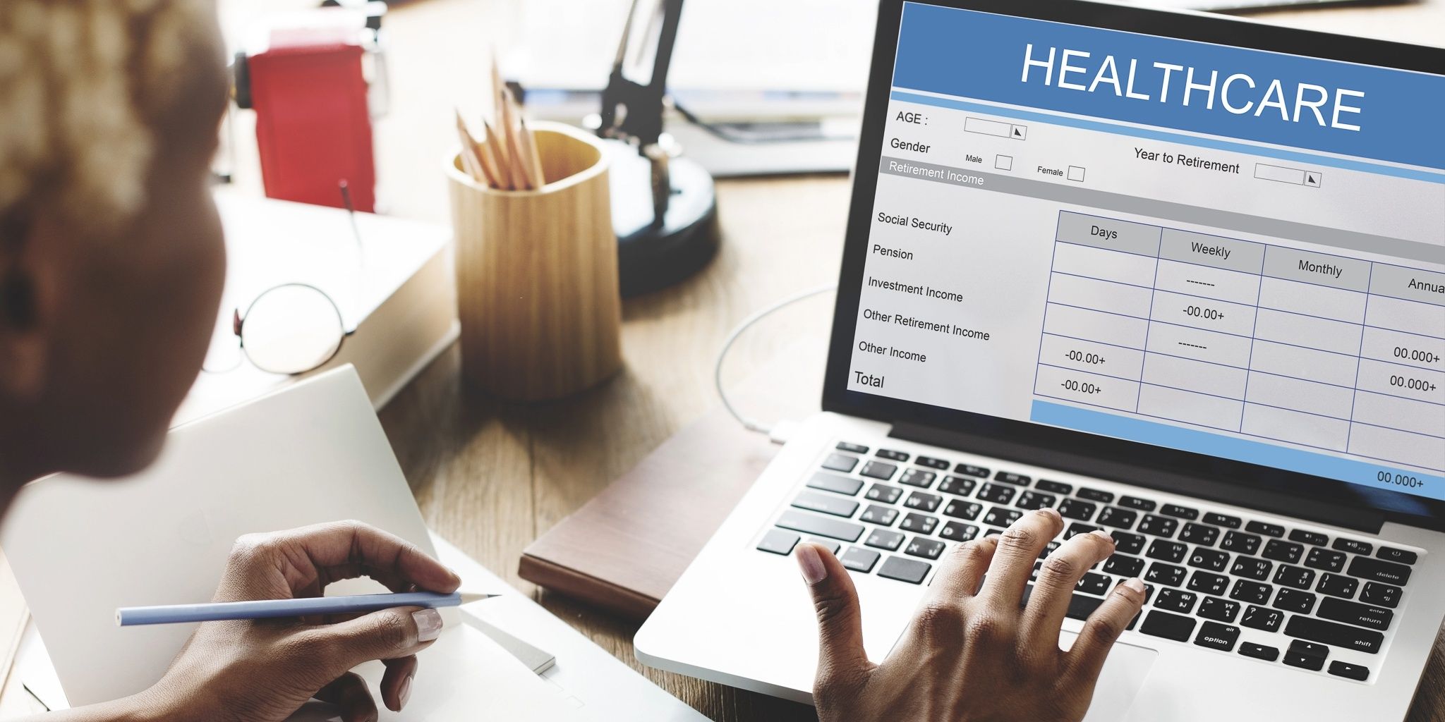 A person using a laptop showing a healthcare financial planning form, with columns for social security, pension, and other income. The person is holding a pen and notebook, working at a desk.