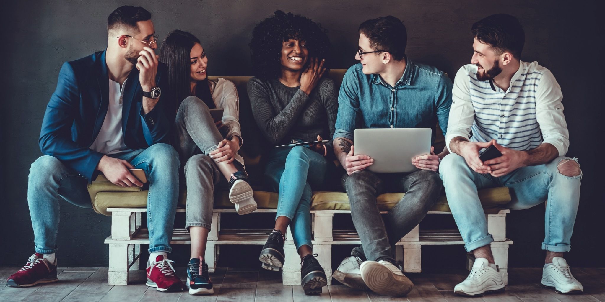 Five young adults sit on a couch, smiling and talking. One uses a laptop, another holds a phone, and they appear relaxed and friendly, dressed casually against a dark background.