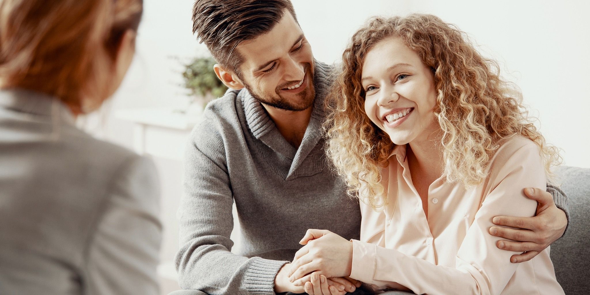 A smiling couple sits close together, holding hands and looking at a woman with her back to the camera, possibly during a counseling or therapy session in a bright, comfortable room.
