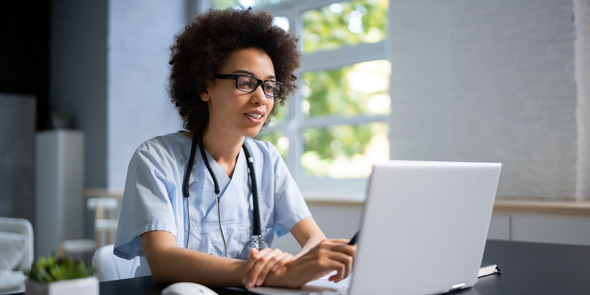A healthcare professional wearing scrubs and a stethoscope sits at a desk, looking at a laptop screen, with natural light coming through large windows in the background.