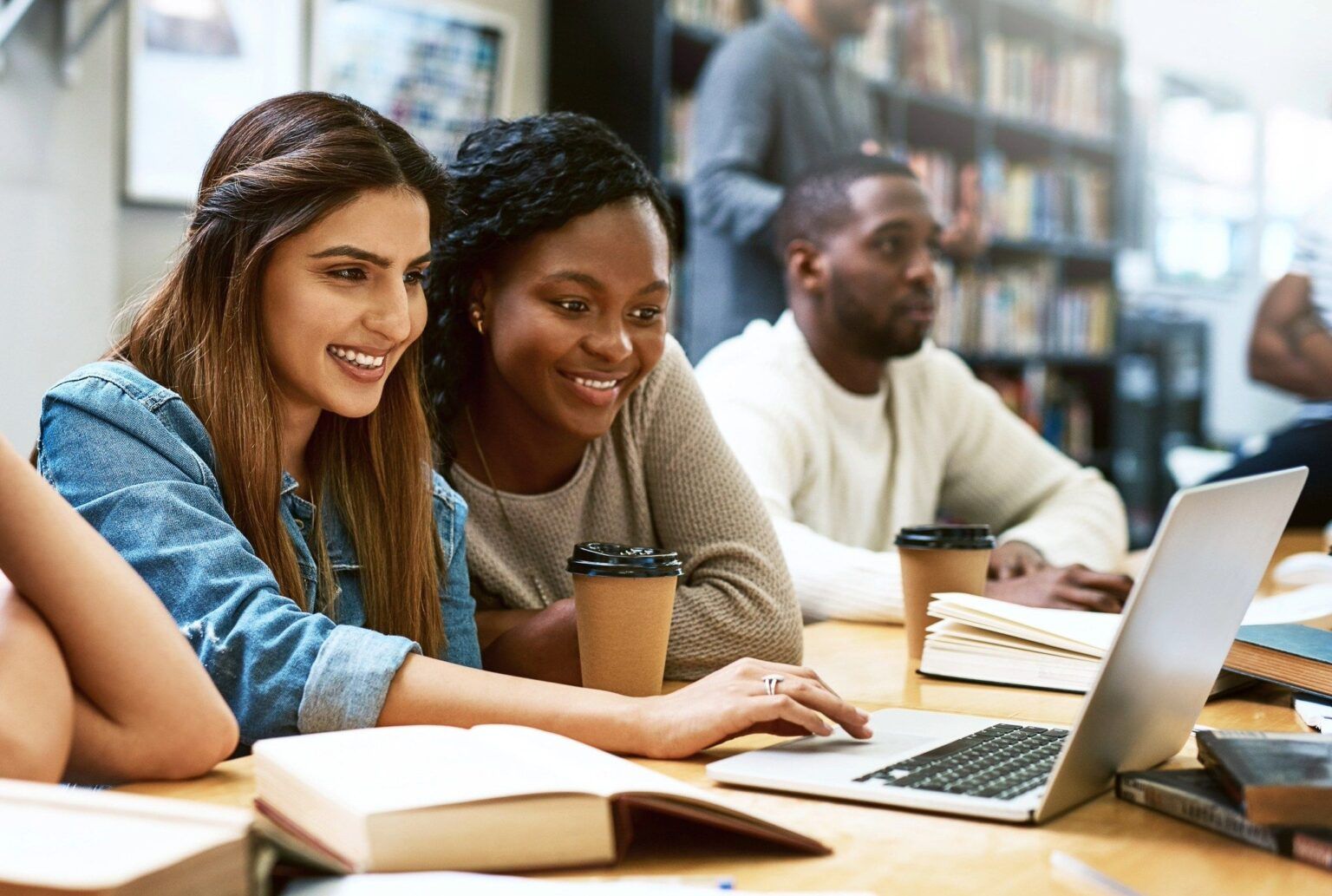 Two young women sitting at a table in a library, smiling and looking at a laptop screen together, with coffee cups and open books in front of them. A man sits in the background, also at the table.