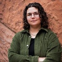 Person with curly brown hair and glasses, wearing a green corduroy jacket over a black shirt, stands with arms crossed in front of a textured red stone wall.