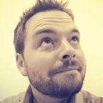 A man with short hair and a beard looks upward with a thoughtful expression, his lips slightly pursed. The image is in sepia tones, giving it a warm, vintage feel.